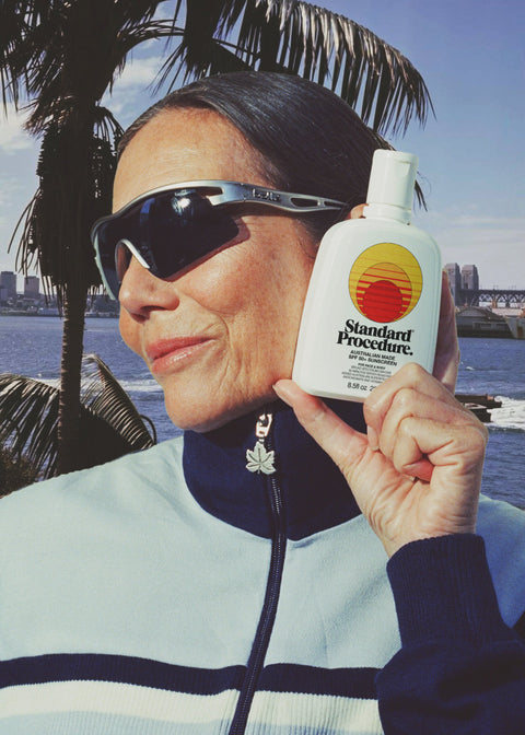 Person holding a bottle of Standard Pacific sunscreen with palm trees and water in the background