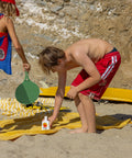 Standard Procedure SPF 50 Sunscreen 250ml bottle on a yellow beach towel with children playing paddle ball in the sand.