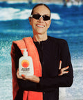 Smiling woman in sunglasses holding a Standard Procedure SPF 50 Sunscreen 500ml pump bottle at the beach.