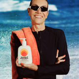 Smiling woman in sunglasses holding a Standard Procedure SPF 50 Sunscreen 500ml pump bottle at the beach.