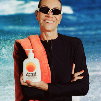 Smiling woman in sunglasses holding a Standard Procedure SPF 50 Sunscreen 500ml pump bottle at the beach.