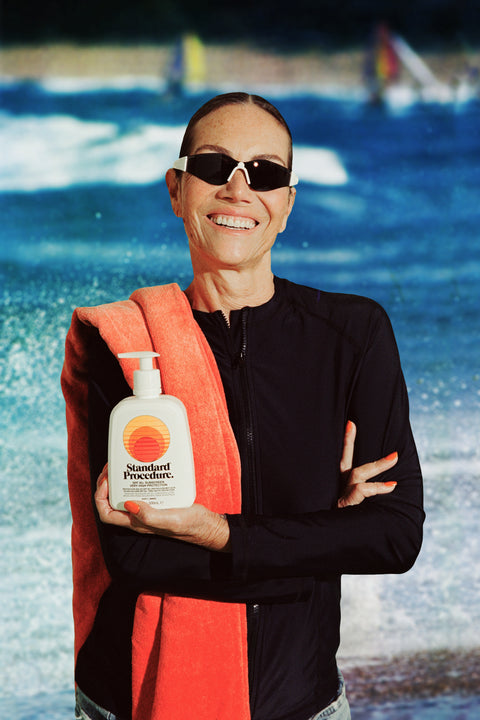 Smiling woman in sunglasses holding a Standard Procedure SPF 50 Sunscreen 500ml pump bottle at the beach.