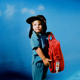 SPF 50 sunscreen attached to a child’s red backpack, with the child wearing a protective surf hat and blue outfit, highlighting portable sun protection for outdoor adventures