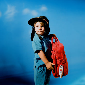SPF 50 sunscreen attached to a child’s red backpack, with the child wearing a protective surf hat and blue outfit, highlighting portable sun protection for outdoor adventures