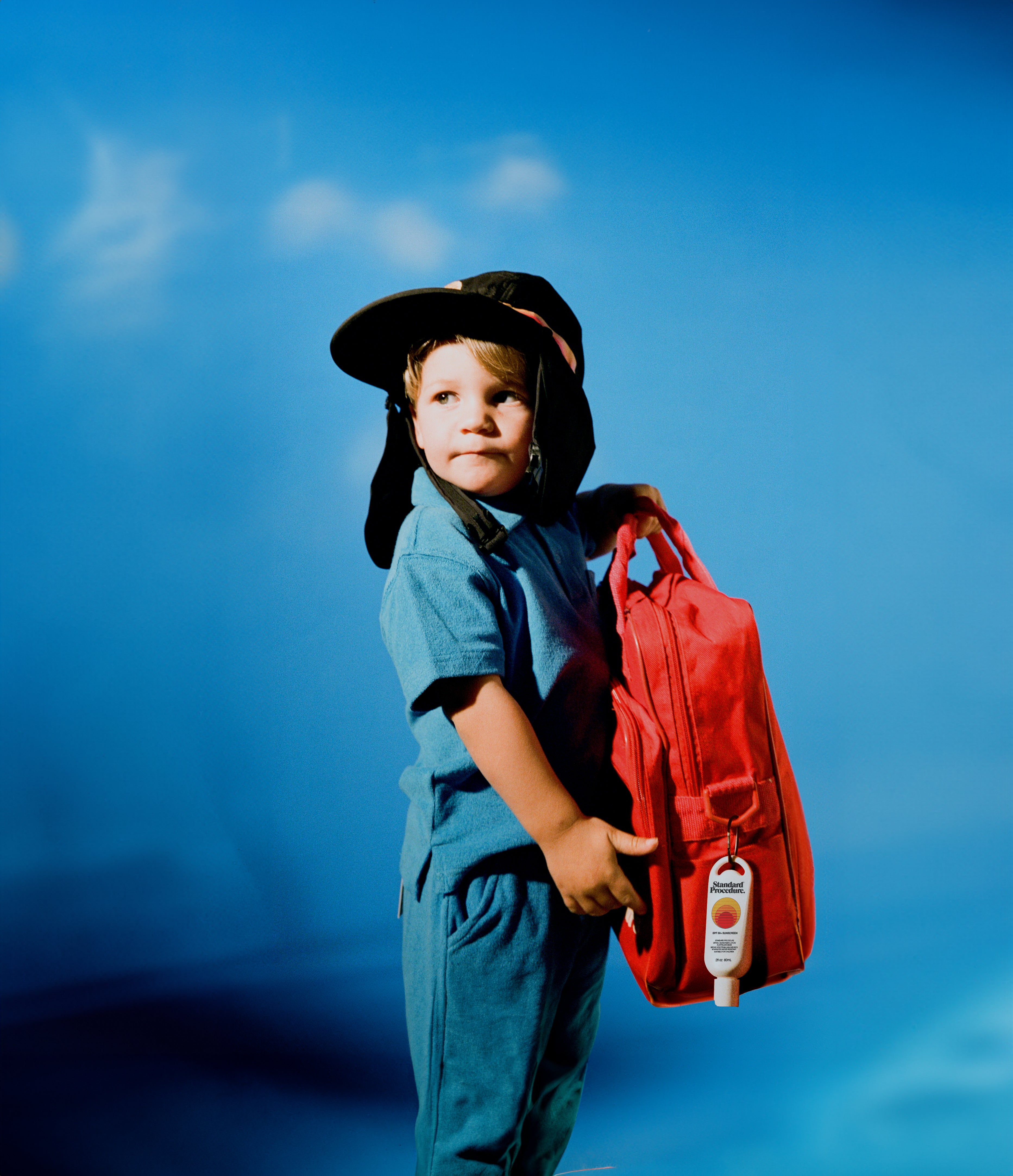 SPF 50 sunscreen attached to a child’s red backpack, with the child wearing a protective surf hat and blue outfit, highlighting portable sun protection for outdoor adventures
