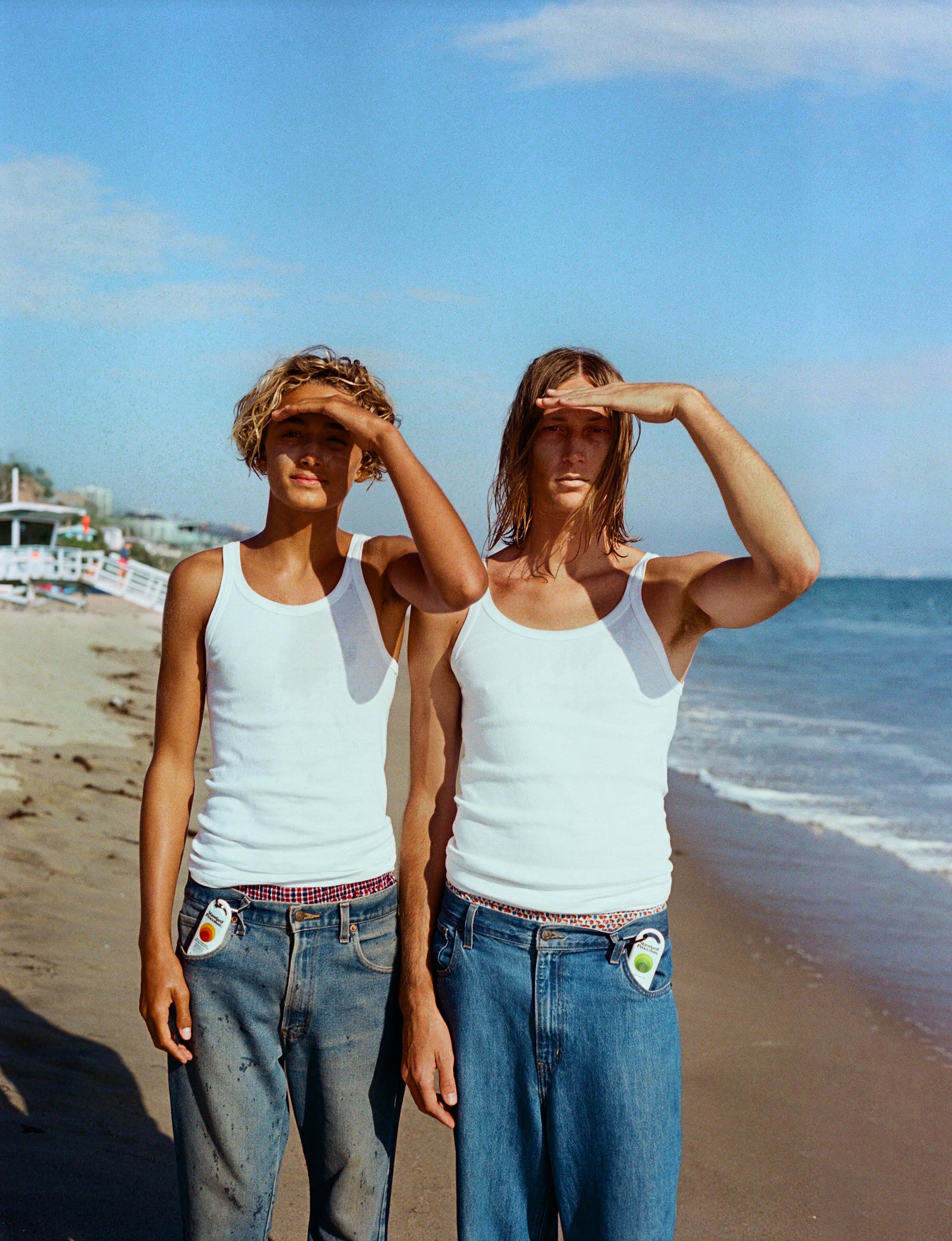 Australian sunscreen and aloe vera clipped to jean pockets as two people in white tank tops shade their eyes on a bright beach
