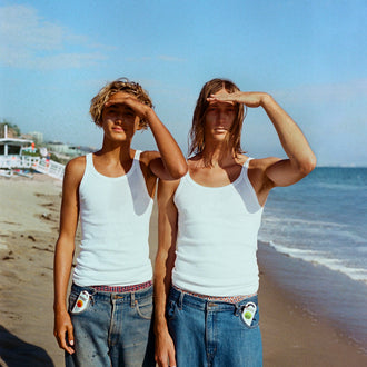 Australian sunscreen and aloe vera clipped to jean pockets as two people in white tank tops shade their eyes on a bright beach
