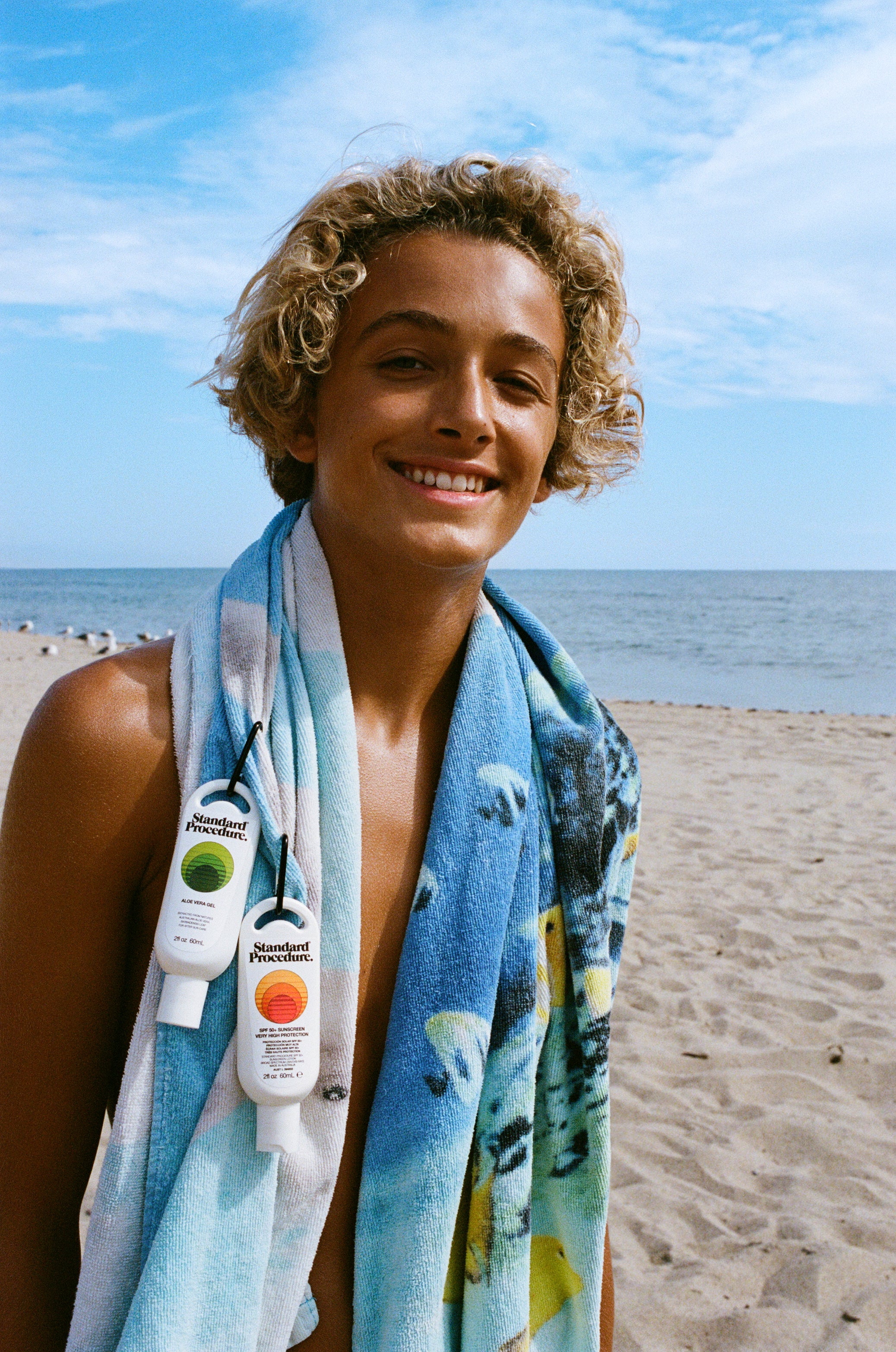 Beachgoer with towel and clip-on Standard Procedure SPF 50 sunscreen and aloe vera gel travel bottles.