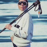 Australian SPF 50 sunscreen clipped to ski poles over a woman in oversized white goggles & a cream sweater standing on snowy slopes

