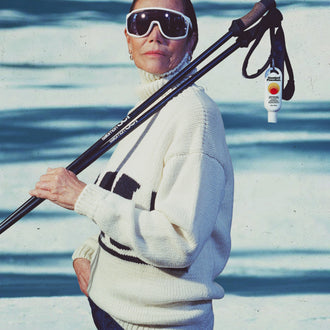 Australian SPF 50 sunscreen clipped to ski poles over a woman in oversized white goggles & a cream sweater standing on snowy slopes
