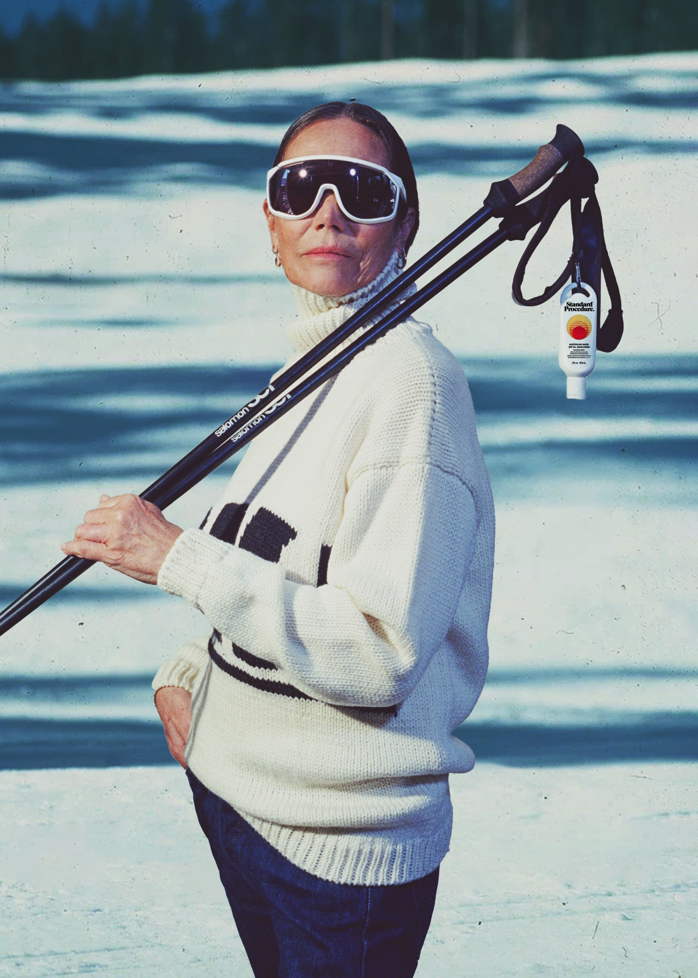 Australian SPF 50 sunscreen clipped to ski poles over a woman in oversized white goggles & a cream sweater standing on snowy slopes
