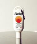 Standard Procedure SPF 50 Sunscreen 60ml travel-size tube with a carabiner clip on a reflective surface.