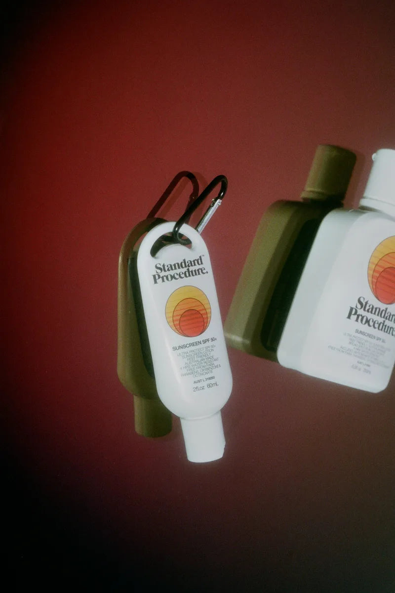 Standard Procedure clip-on SPF 50+ sunscreen mini beside full-size bottle on deep red backdrop.