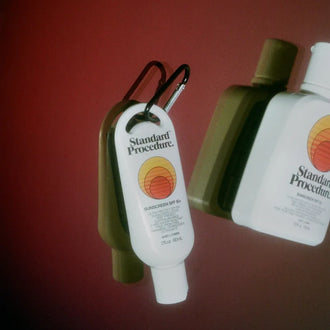 Standard Procedure clip-on SPF 50+ sunscreen mini beside full-size bottle on deep red backdrop.