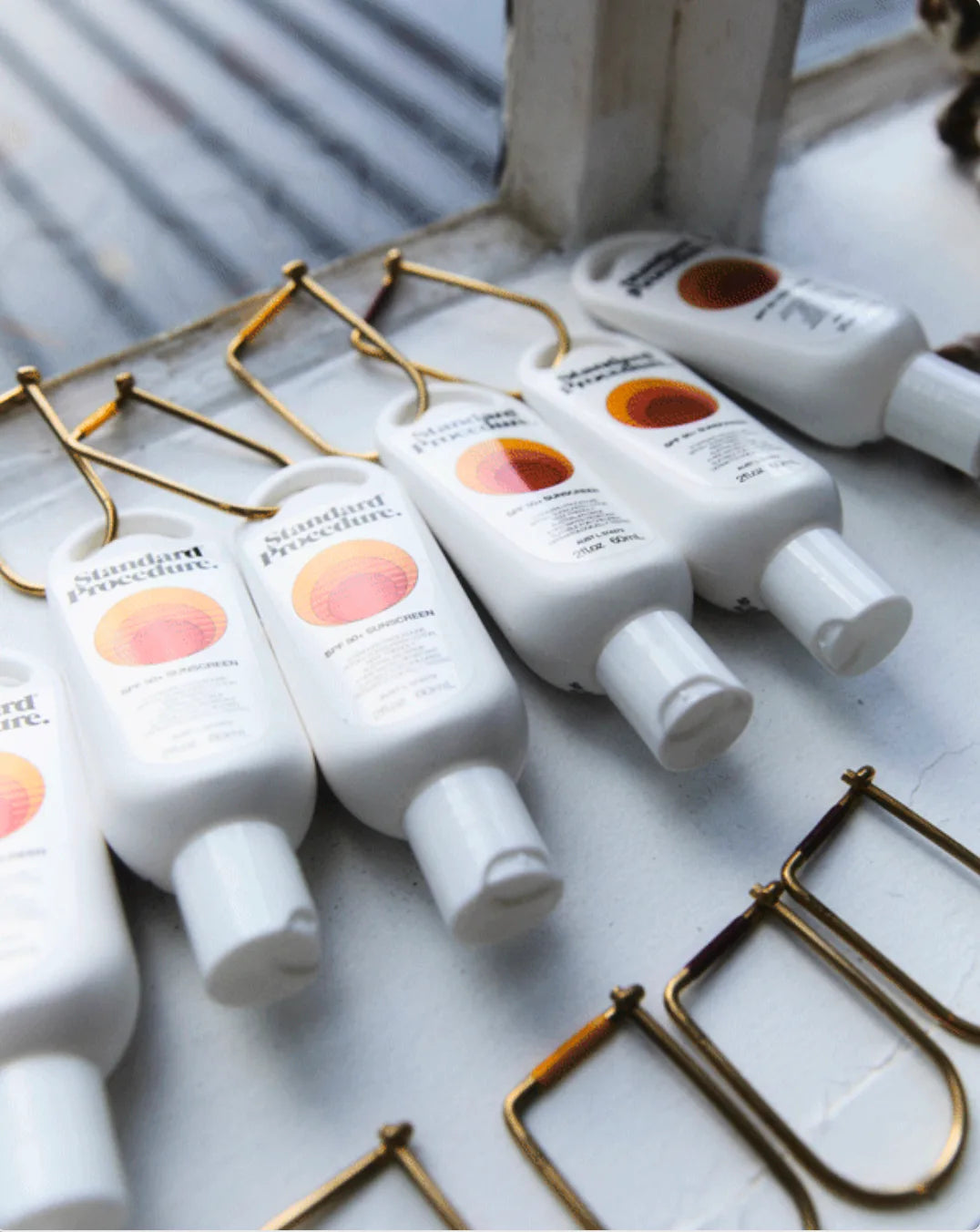 Standard Procedure SPF 50+ sunscreen bottles lined up on a sunlit windowsill with brass letters.