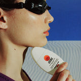 Mineral SPF 50 sunscreen held under a swimmer’s chin in side profile, wearing black goggles against a blue ocean backdrop
