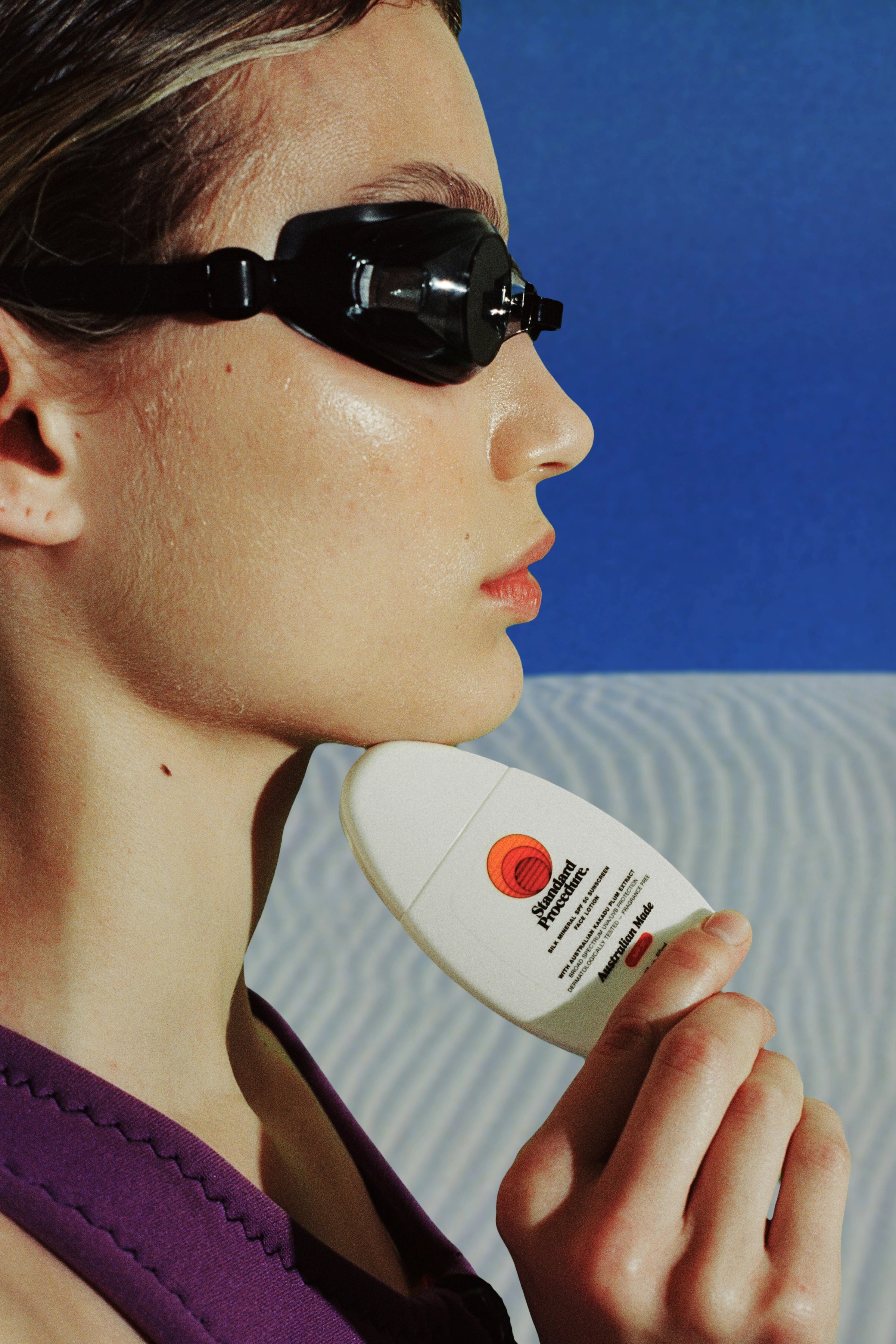 Mineral SPF 50 sunscreen held under a swimmer’s chin in side profile, wearing black goggles against a blue ocean backdrop
