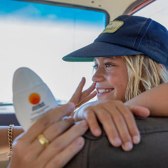 Mineral sunscreen broad spectrum SPF 50 being applied by a smiling woman in a car, wearing a navy cap and gold jewelry, emphasizing everyday sun protection