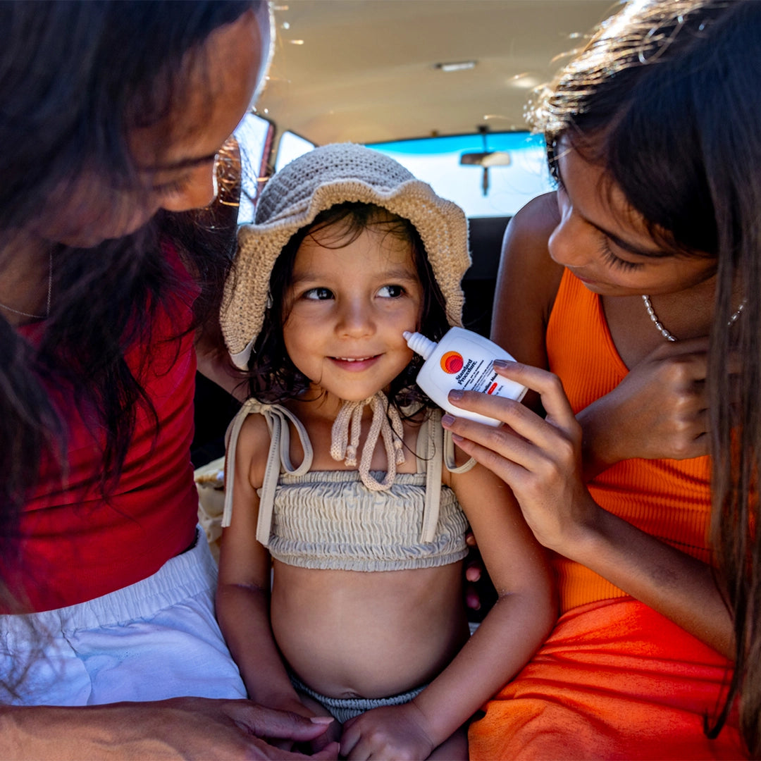 Mineral SPF 50 sunscreen being gently applied to a smiling child’s cheek by an adult, showing sun protection care during a family beach outing