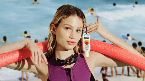 Woman in a colorful swimsuit holding a key chain sunscreen bottle on a beach with people in the background