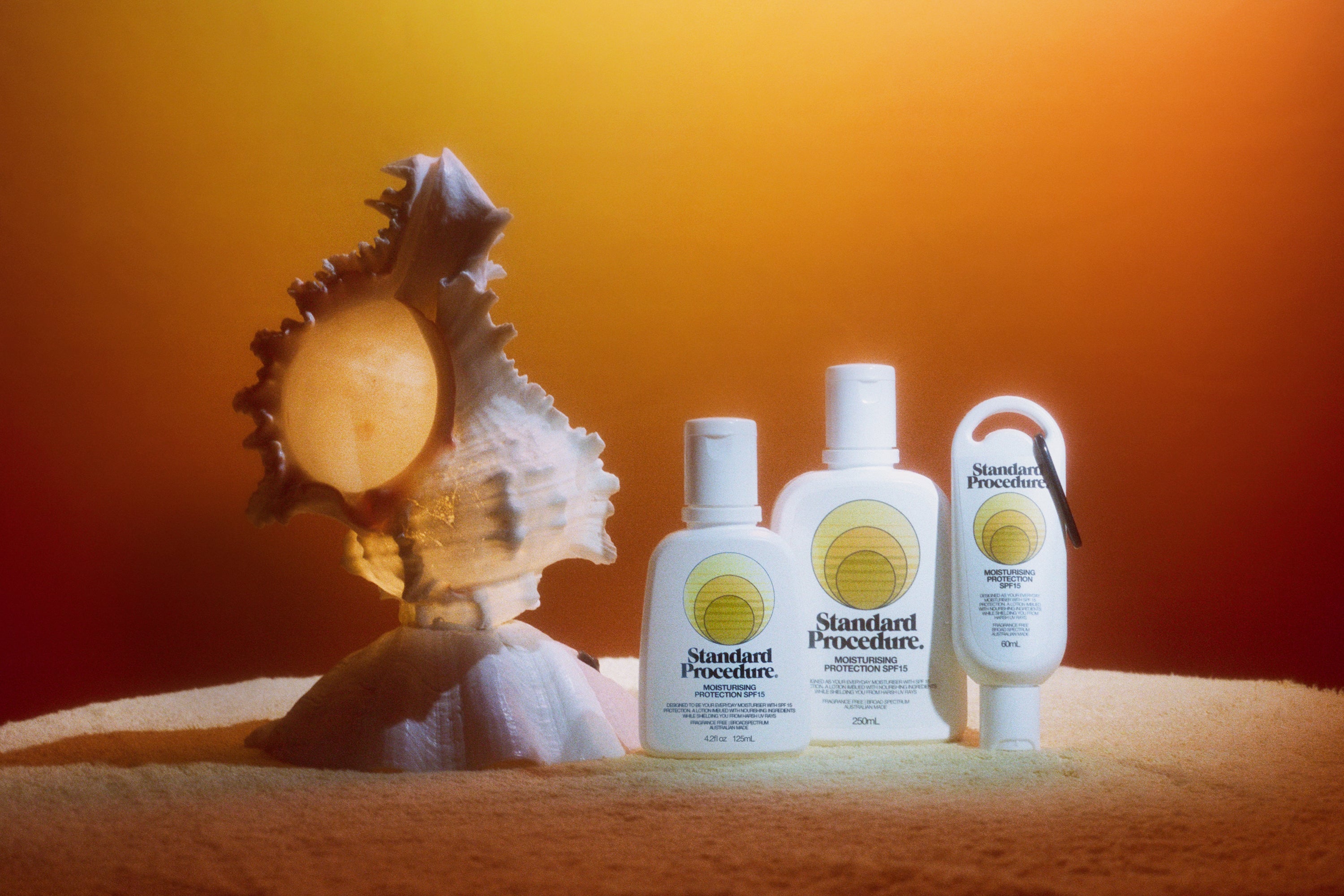 Three Standard Procedure SPF 15 moisturiser bottles beside a seashell on towel in sunset glow.