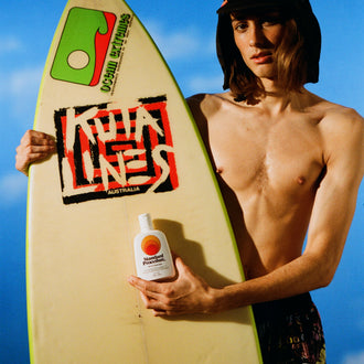 Sun block SPF 50 bottle held by a surfer with a board and wearing a sun-protective hat under a clear blue sky.