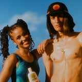 Australian made sunscreen held by a smiling woman next to a man wearing a sun-protective hat under a clear blue sky.