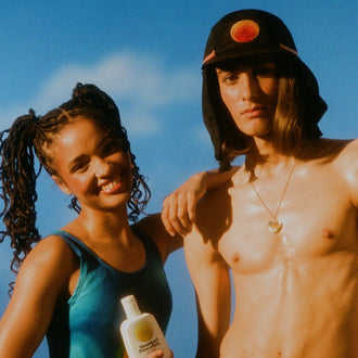Australian made sunscreen held by a smiling woman next to a man wearing a sun-protective hat under a clear blue sky.