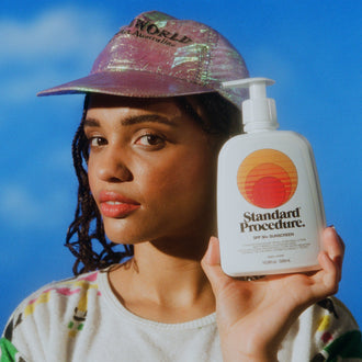 Broad spectrum sunscreen SPF 50 bottle held by a model wearing a pink iridescent cap against a bright blue sky backdrop.