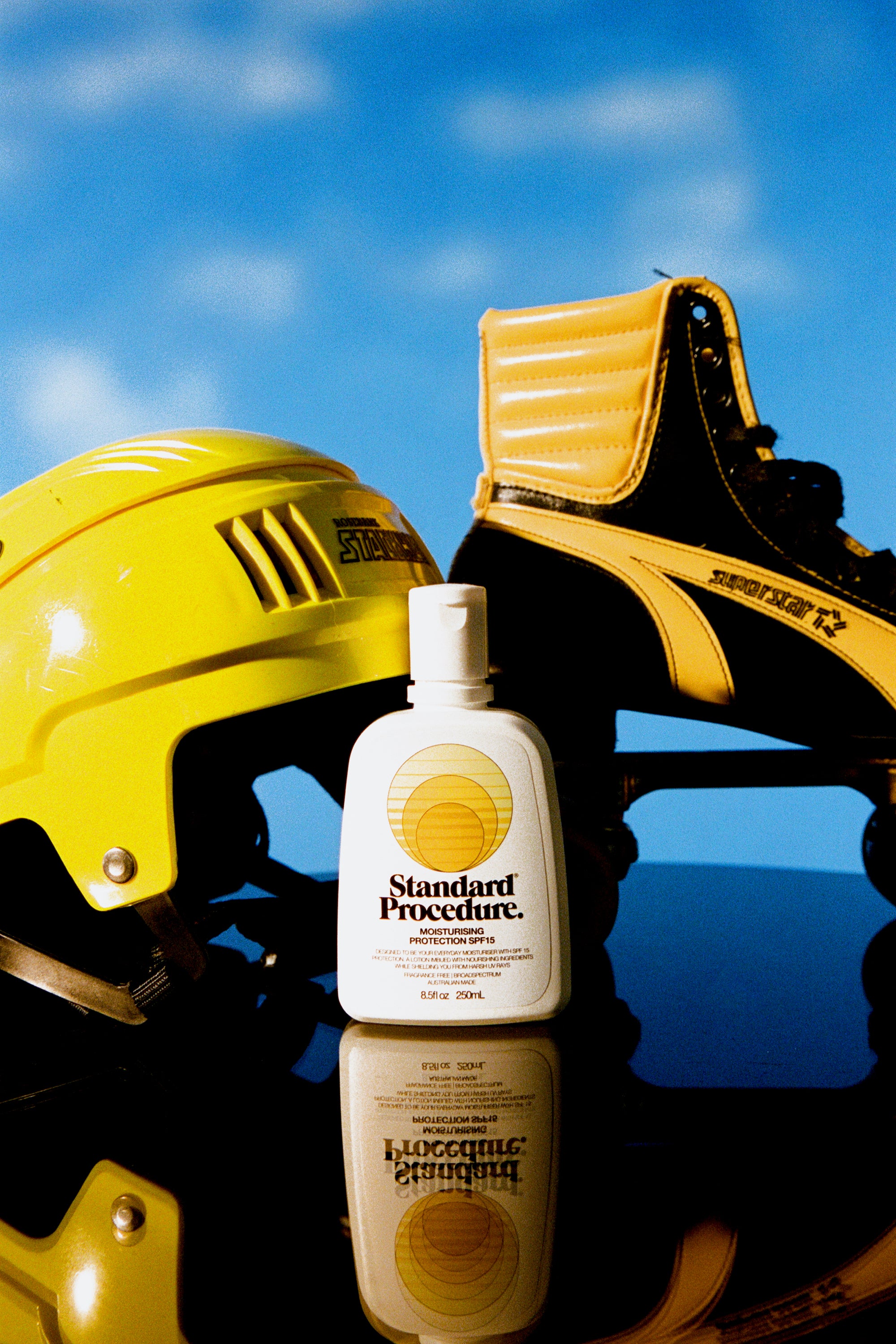 Standard Procedure SPF 15 moisturising sunscreen bottle beside yellow helmet and skate against blue sky.