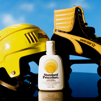 Standard Procedure SPF 15 moisturising sunscreen bottle beside yellow helmet and skate against blue sky.