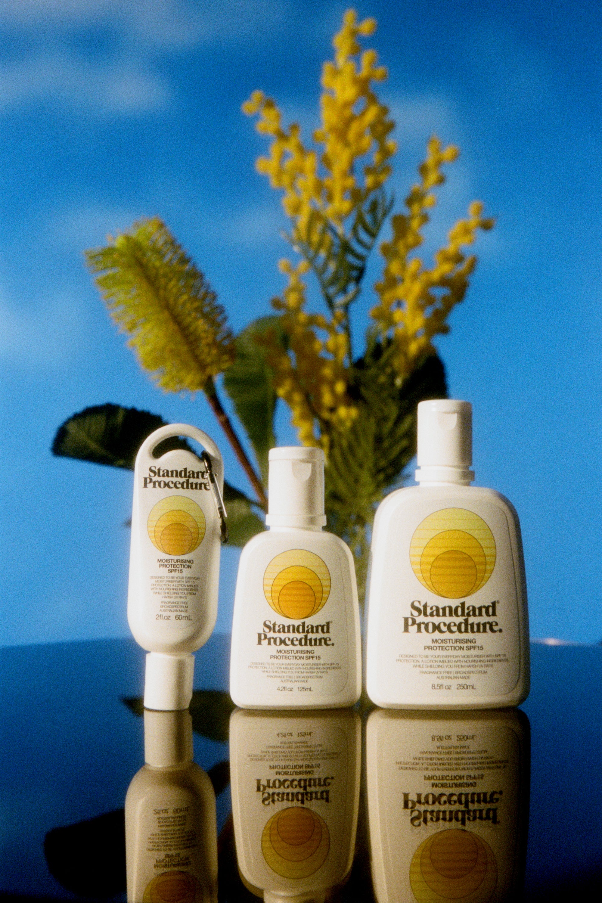 Three Standard Procedure SPF 15 sunscreen bottles with wattle sprigs against a blue sky backdrop.