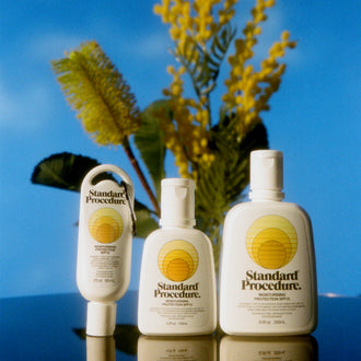 Three Standard Procedure SPF 15 sunscreen bottles with wattle sprigs against a blue sky backdrop.