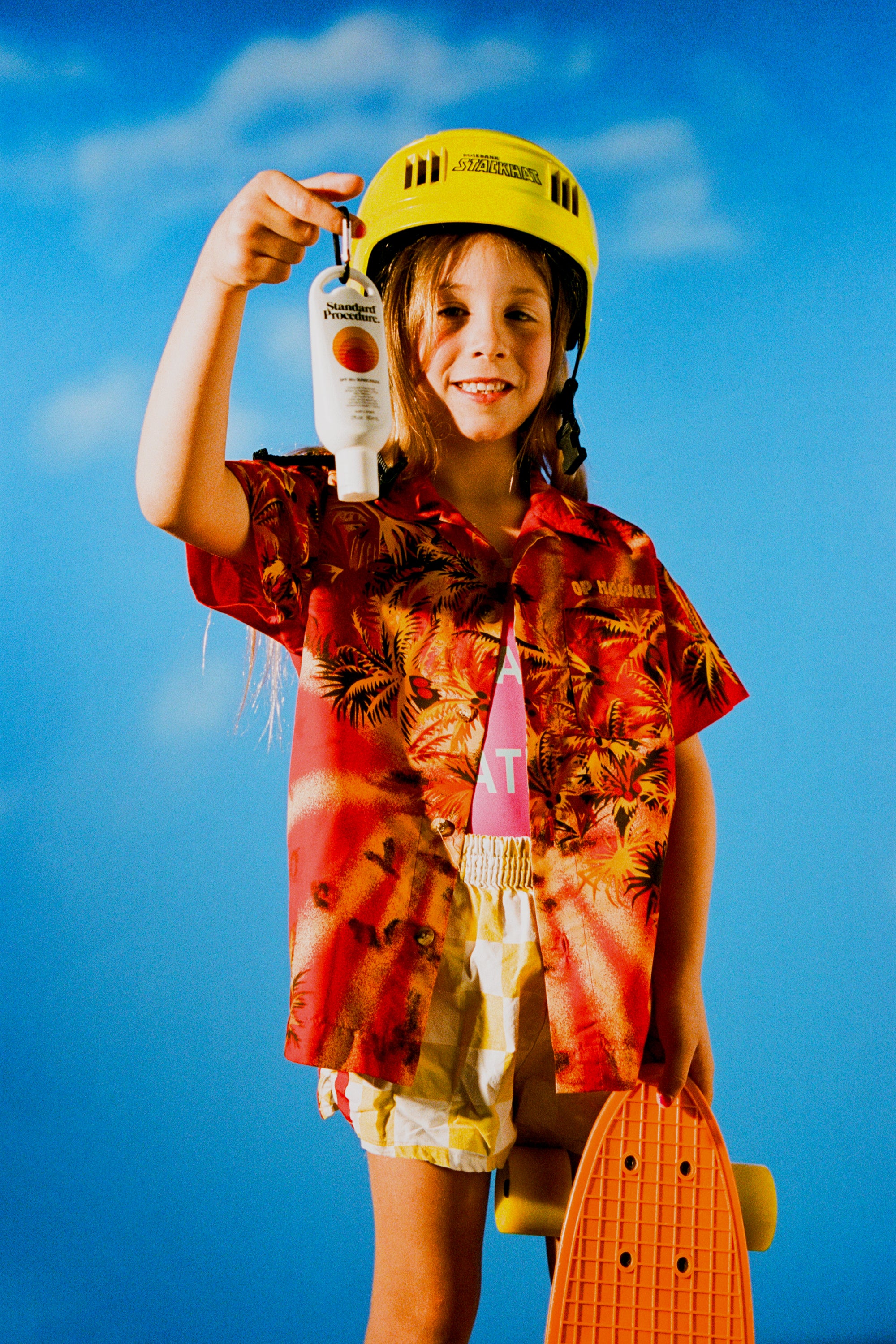 Sun Protect SPF 50 held by a smiling child in a tropical shirt and helmet with a skateboard against a blue sky background.