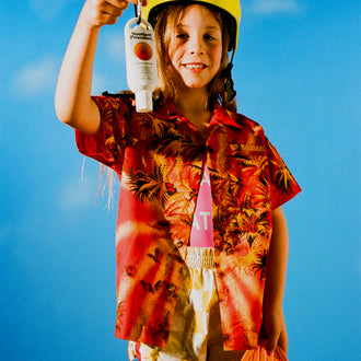 Sun Protect SPF 50 held by a smiling child in a tropical shirt and helmet with a skateboard against a blue sky background.