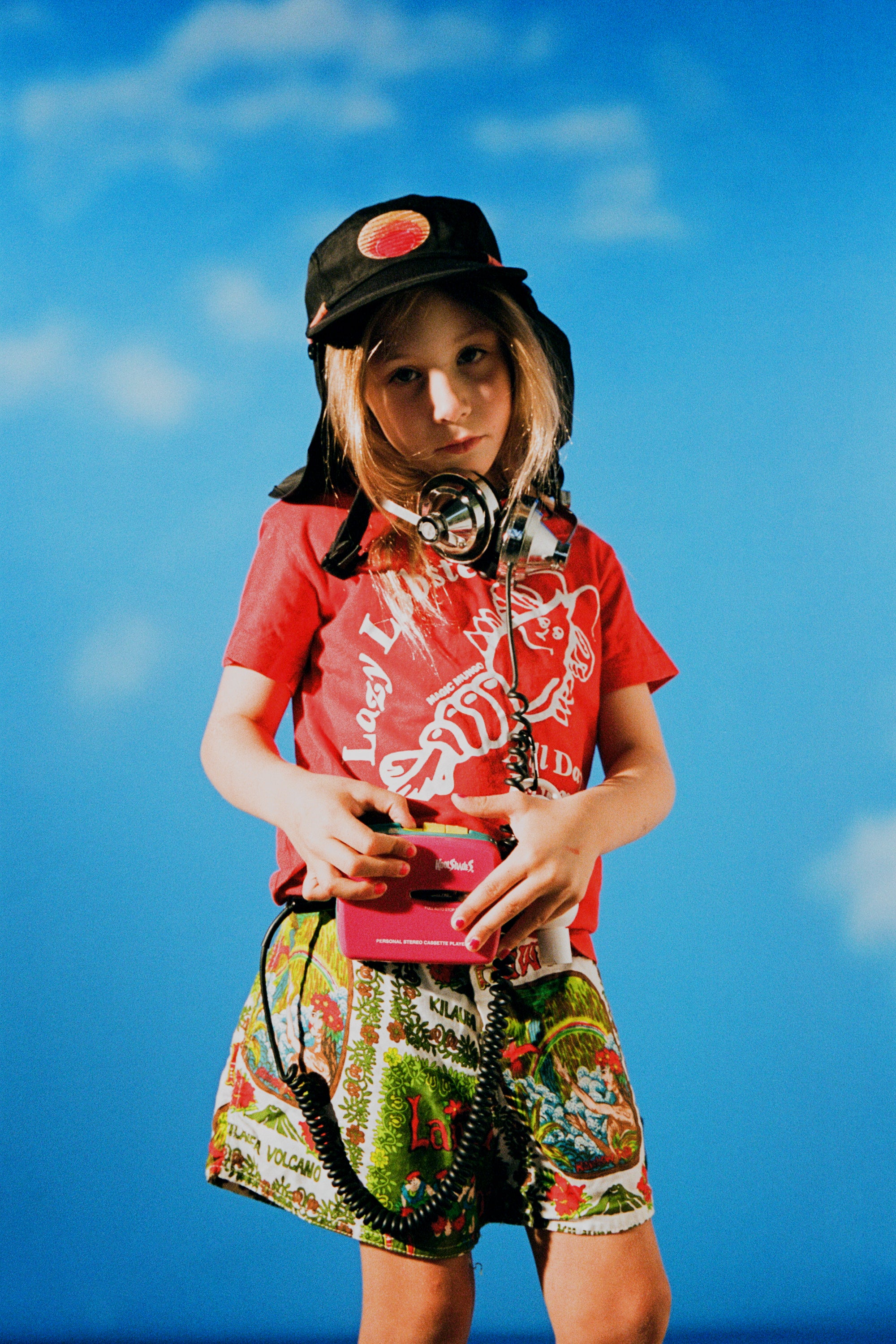 Vitamin C with sunscreen not shown, but child wearing a tropical outfit and sun hat listens to music against a blue sky backdrop.