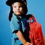 Child wearing black kids surf hat with neck flap, holding red bag against blue sky.