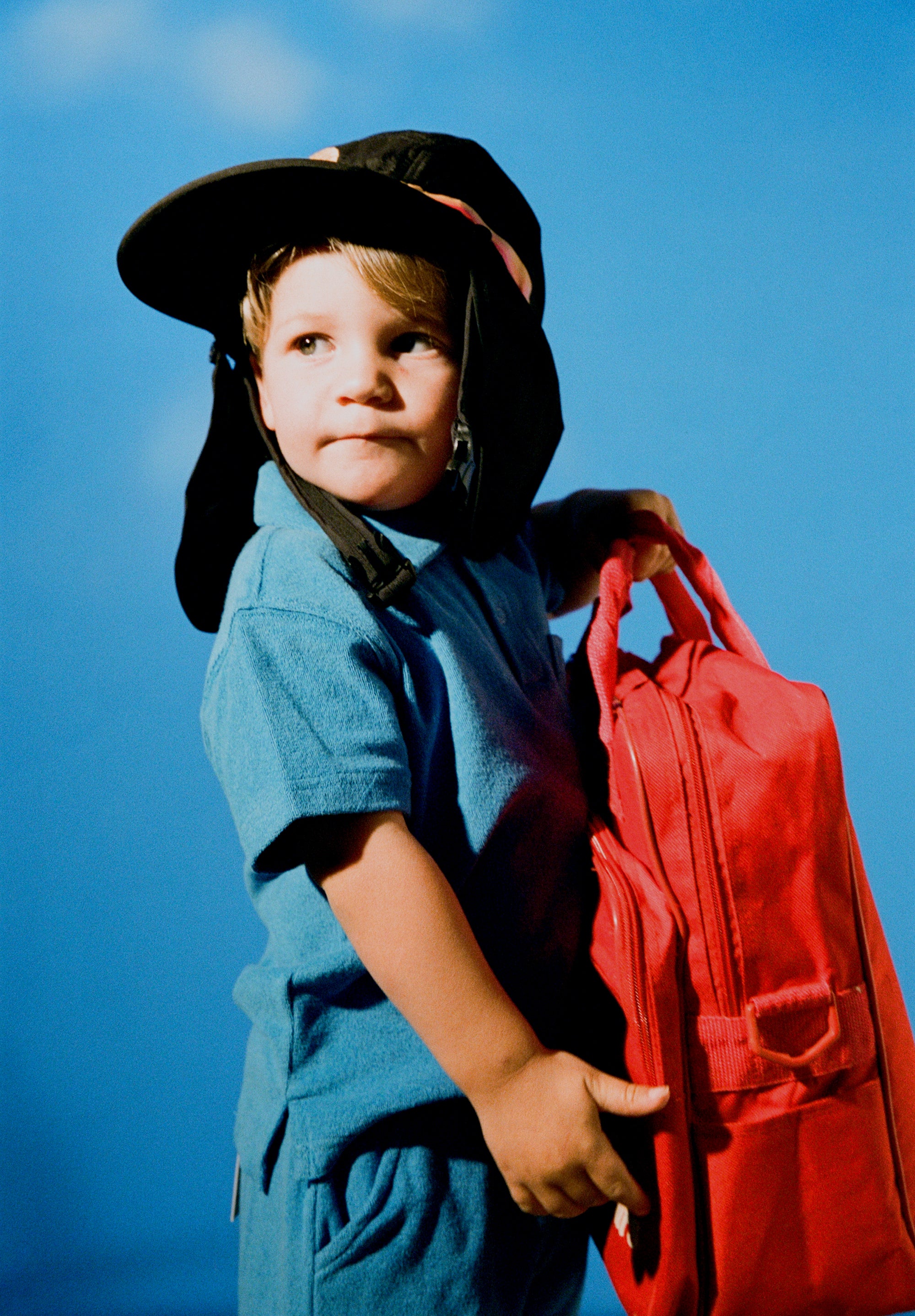 Child wearing black kids surf hat with neck flap, holding red bag against blue sky.