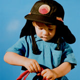 Child wearing the black Grom Pack kids surf hat with neck flap against a blue sky.