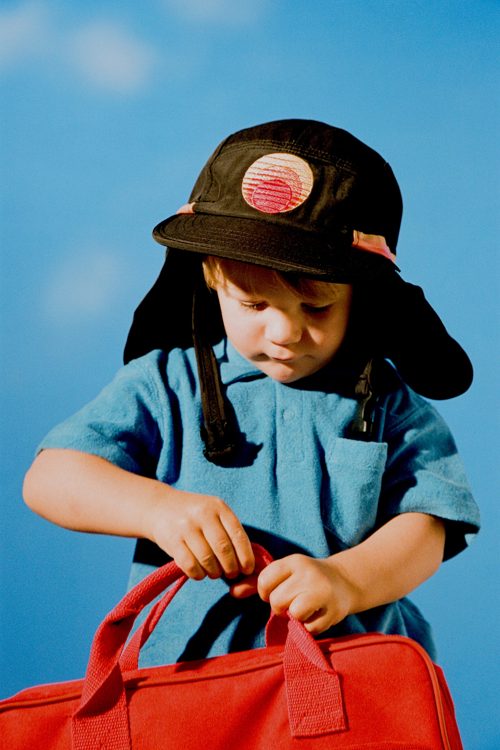 Child wearing the black Grom Pack kids surf hat with neck flap against a blue sky.