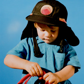 Child wearing the black Grom Pack kids surf hat with neck flap against a blue sky.