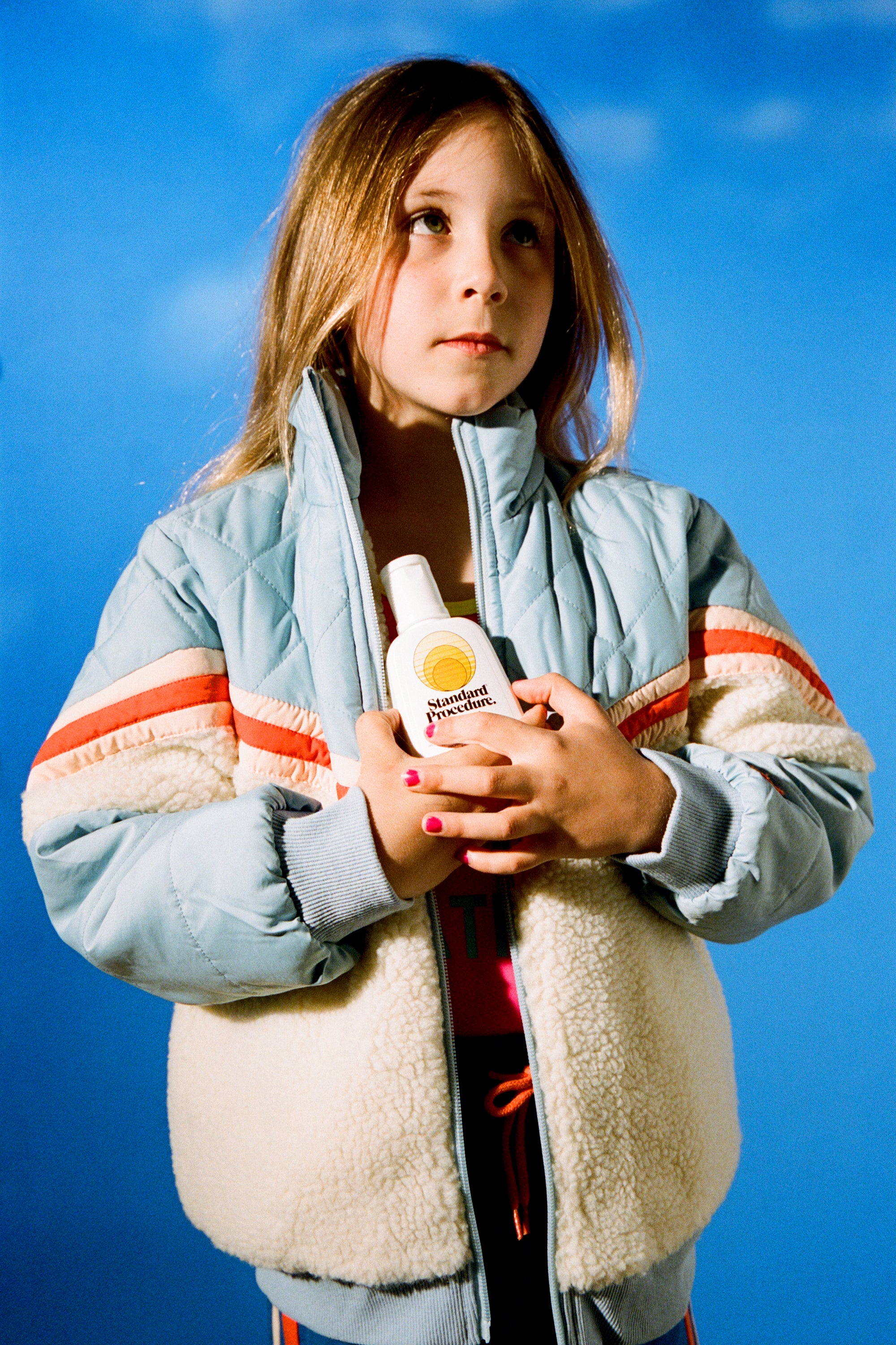 Child in retro blue jacket holding Standard Procedure SPF 15 sunscreen against a sky backdrop.