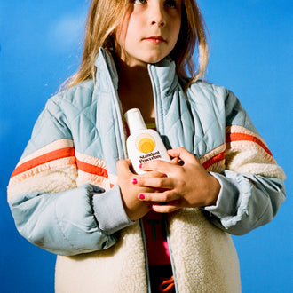 Child in retro blue jacket holding Standard Procedure SPF 15 sunscreen against a sky backdrop.