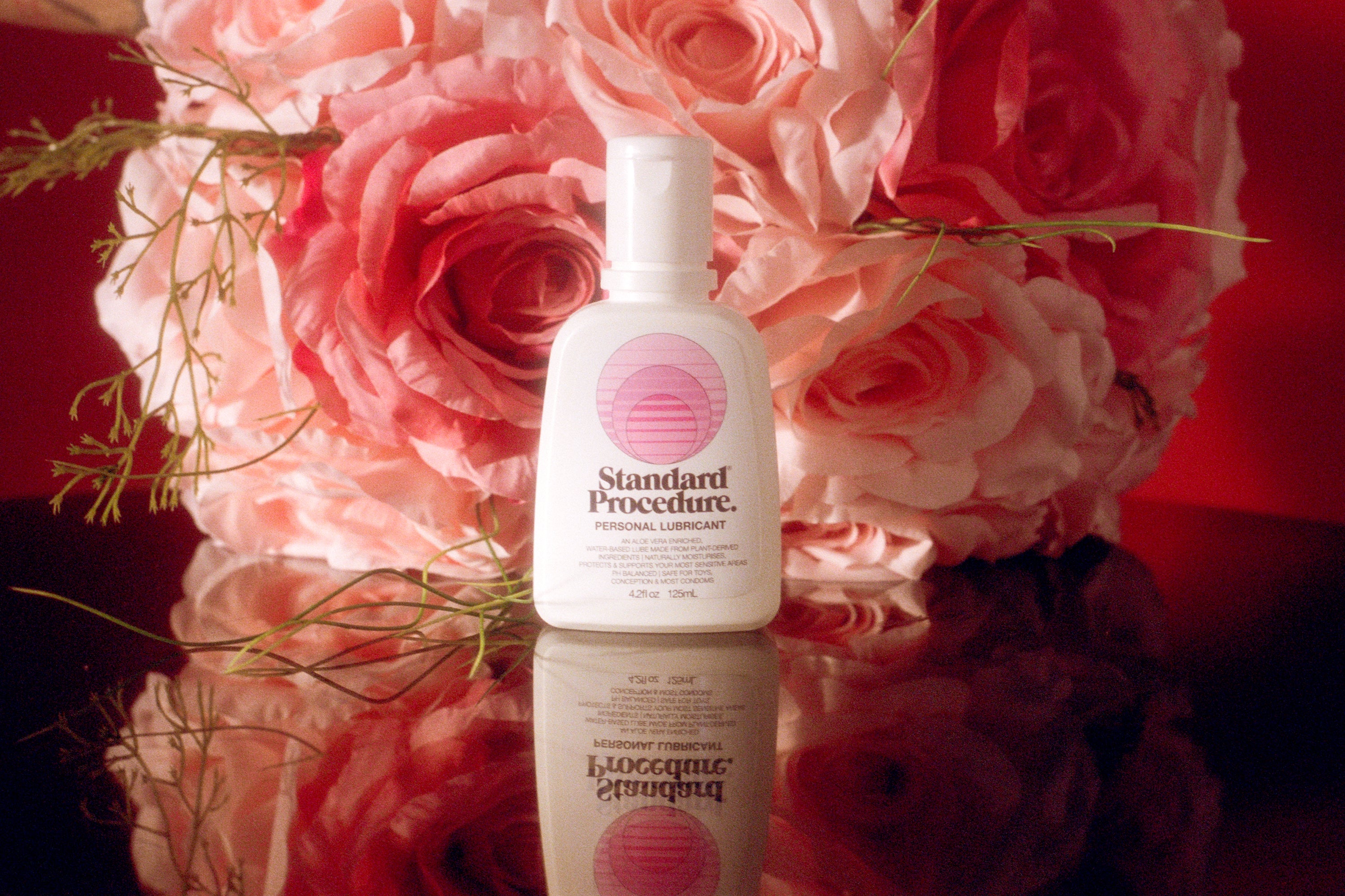 Pleasure Jelly aloe vera lubricant bottle styled against pink roses on a reflective surface.