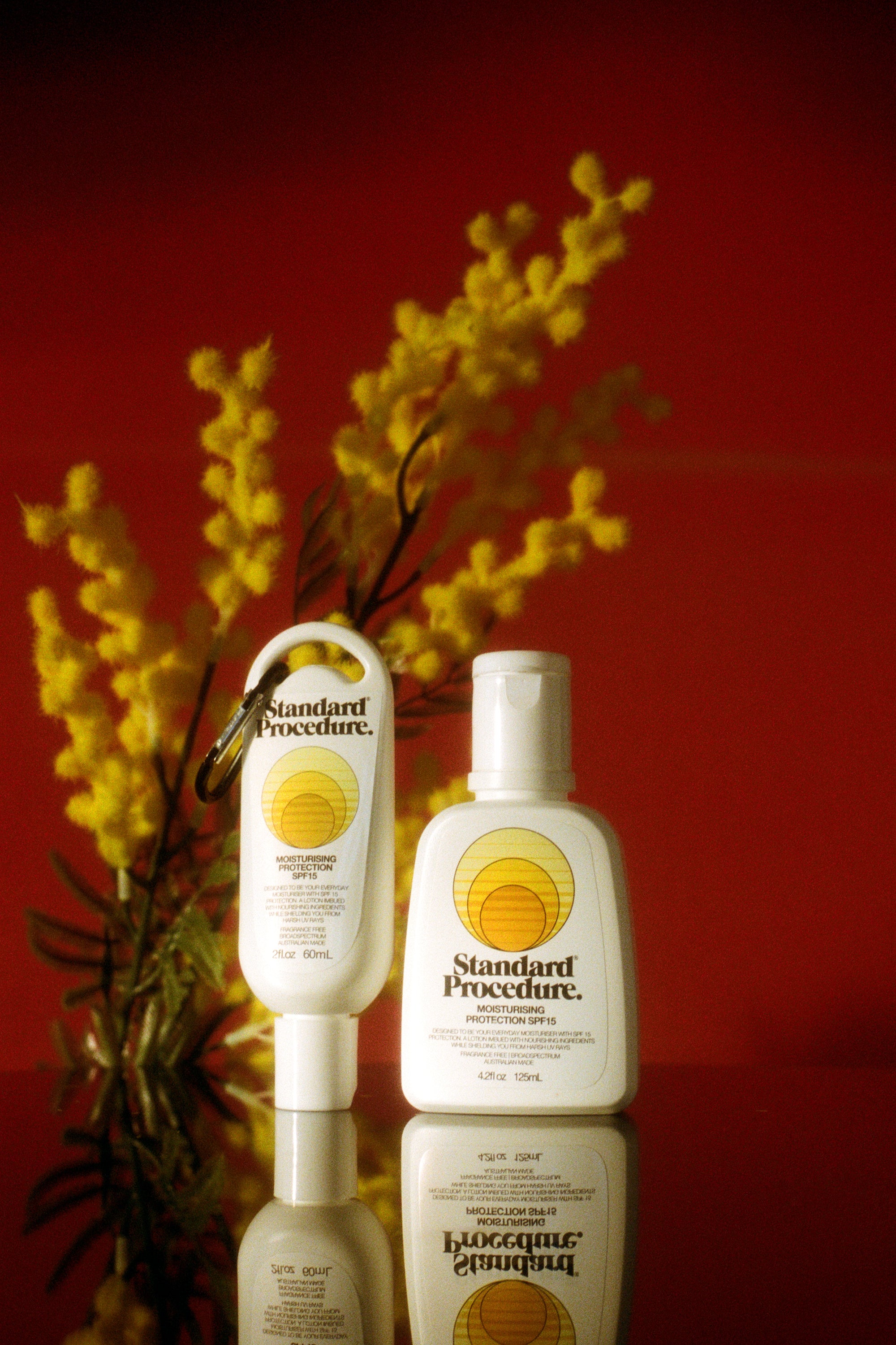 Standard Procedure SPF 15 moisturiser bottles styled with golden wattle against a deep red backdrop.