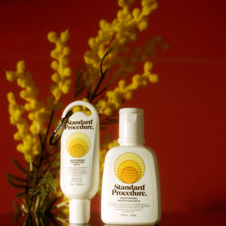 Standard Procedure SPF 15 moisturiser bottles styled with golden wattle against a deep red backdrop.