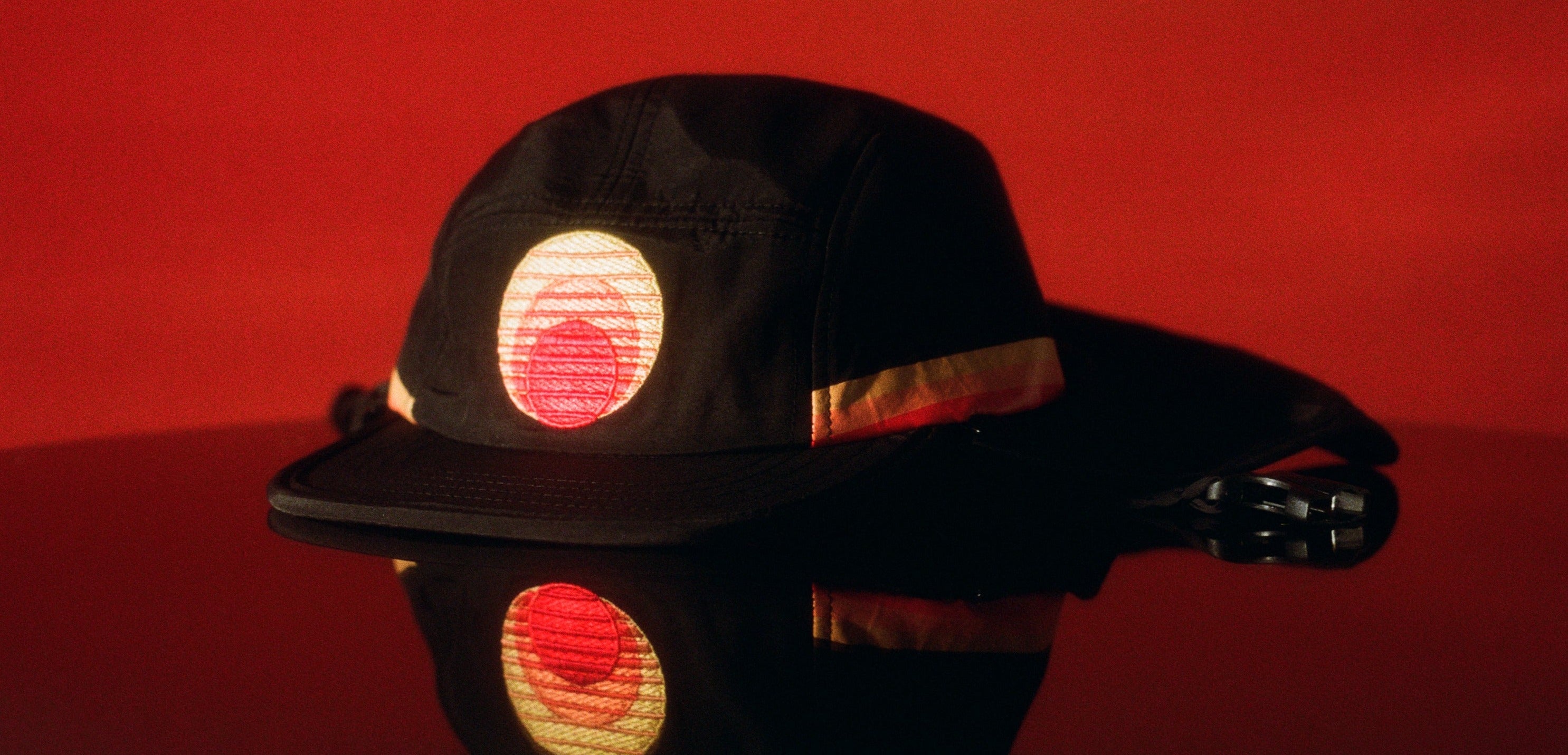 Black kids surf hat with embroidered sunset logo, reflected on a glossy red surface.