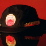 Black kids surf hat with embroidered sunset logo, reflected on a glossy red surface.