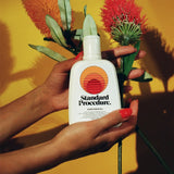 Vitamin C with sunscreen bottle held by hands alongside red and orange flowers against yellow backdrop.