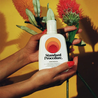 Vitamin C with sunscreen bottle held by hands alongside red and orange flowers against yellow backdrop.